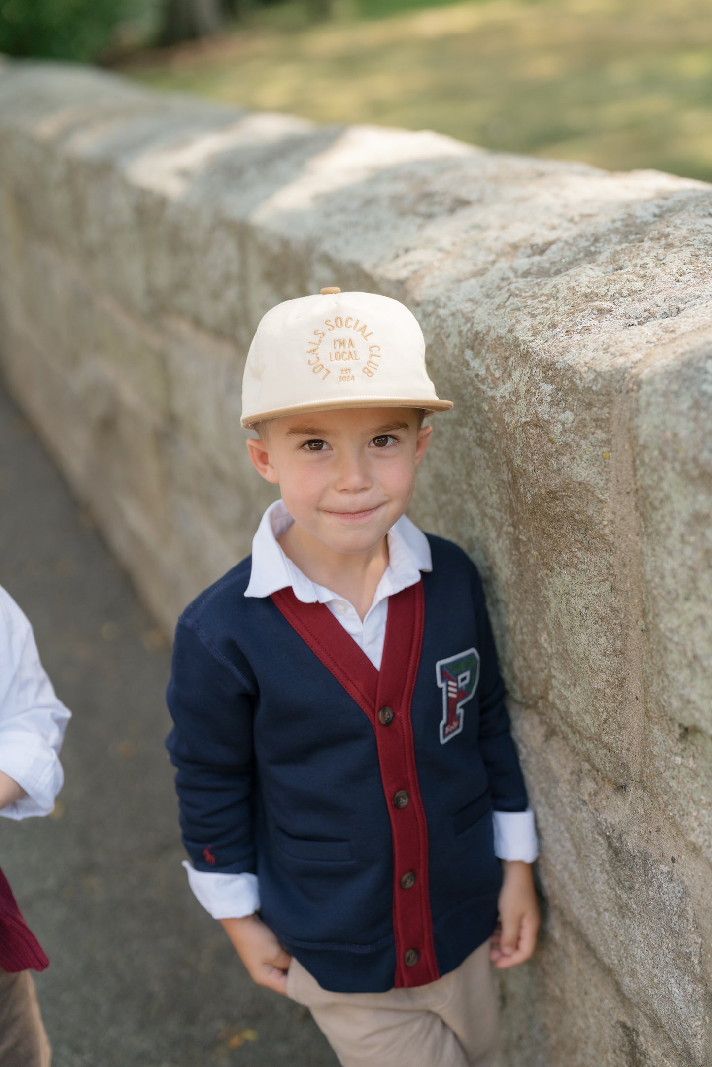 Young boy in a formal outfit with a cap standing against a stone wall.